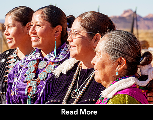 4 indigenous women in tribal attire look out smiling at a sunny mountain landscape